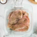 Raw chicken breasts seasoned with herbs and pepper are arranged in a glass baking dish on a marble countertop, surrounded by sliced mozzarella, salt, pepper, and fresh basil.