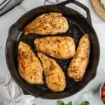 Five cooked chicken breasts are arranged in a cast iron skillet on a marble countertop, with tongs, a cloth, fresh basil, and a small bowl of tomatoes nearby.