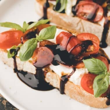 Slices of caprese garlic bread topped with mozzarella, cherry tomatoes, basil leaves, and a drizzle of balsamic glaze on a white plate.