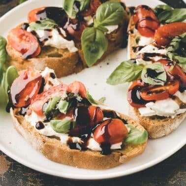 Three slices of caprese garlic bread topped with mozzarella, cherry tomatoes, fresh basil, and balsamic glaze on a white plate.