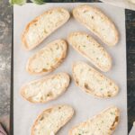 Eight slices of bread topped with herb butter are arranged on parchment paper with a butter knife and a partially unwrapped stick of herb butter nearby.