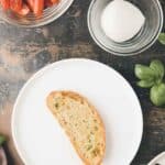 A slice of toasted bread on a white plate, with bowls of cherry tomatoes and mozzarella, and fresh basil leaves nearby on a dark surface.