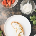 A slice of bread with white cheese on a white plate, next to bowls of cherry tomatoes and cheese, and fresh basil leaves on a dark surface.