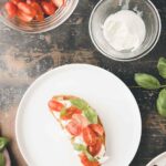 A slice of bread topped with ricotta, cherry tomato halves, and basil sits on a white plate beside bowls of tomatoes, ricotta, fresh basil, and a drizzle of balsamic glaze.