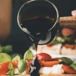 A hand pours balsamic glaze from a small glass pitcher onto a slice of caprese garlic bread topped with mozzarella, tomatoes, and fresh basil.
