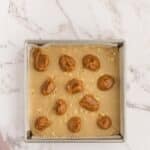 A square baking pan filled with light brown batter, topped with dollops of caramel sauce, sits on a marble countertop.