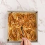 A hand uses a spoon to swirl caramel into batter in a square baking pan on a marble countertop.