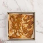 A square baking pan with a caramel-swirl cake on a white marble surface, partially covered by a light purple cloth in the corner.