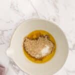 A mixing bowl containing melted butter, brown sugar, and granulated sugar sits on a marble countertop next to a pink cloth.