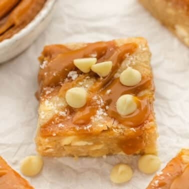 A caramel blondie topped with white chocolate chips and a drizzle of caramel sauce, placed on crumpled parchment paper.