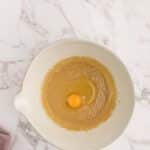 A mixing bowl with light brown batter and a raw egg in the center, placed on a white marble surface next to a folded mauve cloth.