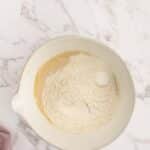 A mixing bowl containing flour, sugar, and a whole egg sits on a marble surface next to a folded pink cloth.