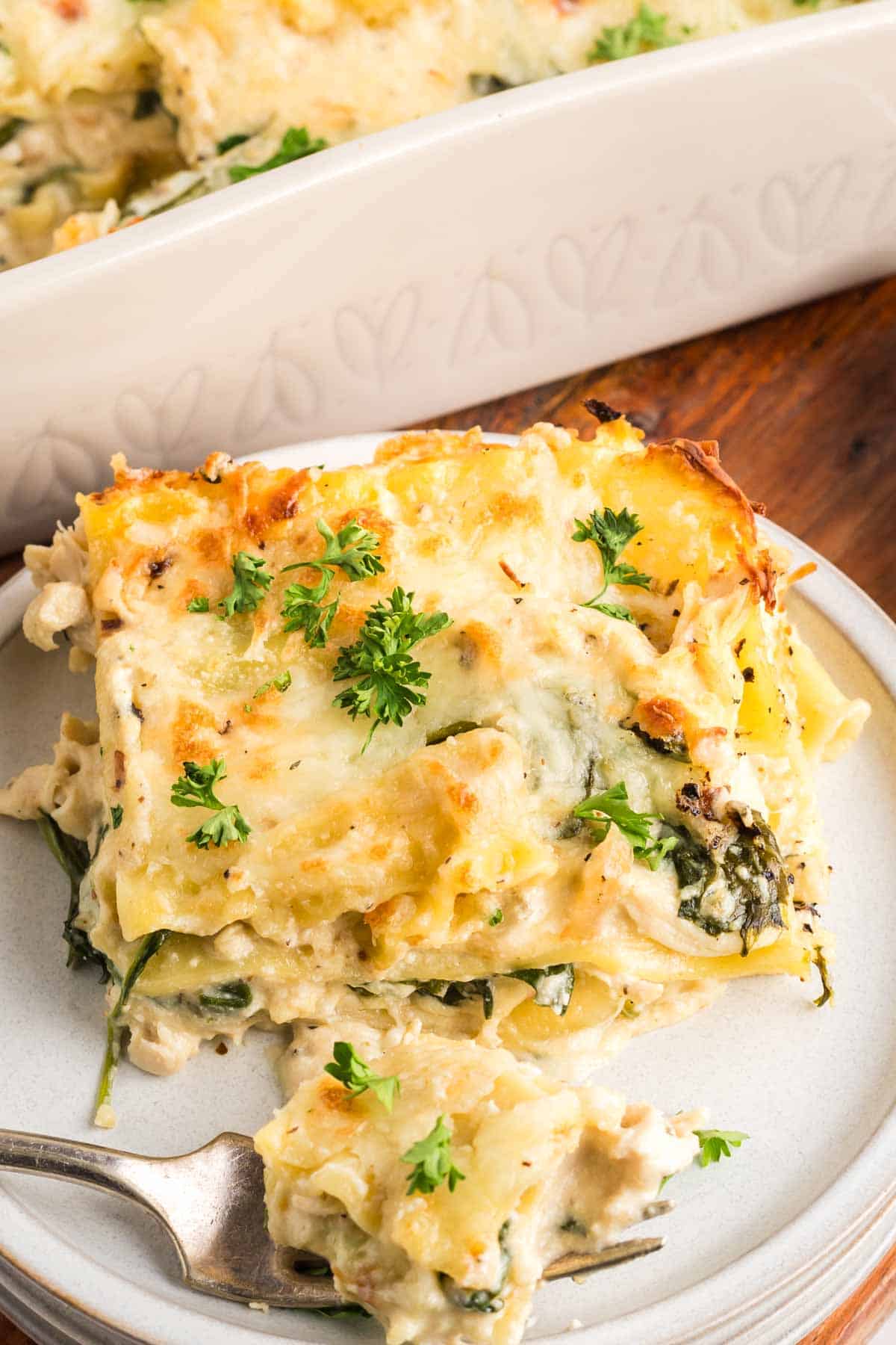 A serving of chicken alfredo lasagna with spinach and cheese is plated, garnished with fresh parsley, with a fork and baking dish in the background.