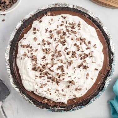 A no bake chocolate pie with a whipped cream topping and chocolate shavings, in a foil pie pan on a white surface next to a pie server and a blue napkin.