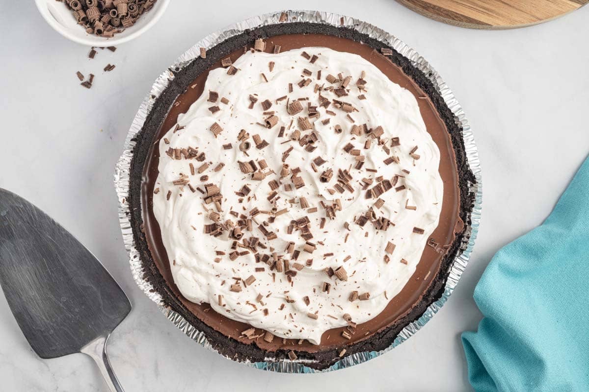 A no bake chocolate pie with a whipped cream topping and chocolate shavings, in a foil pie pan on a white surface next to a pie server and a blue napkin.