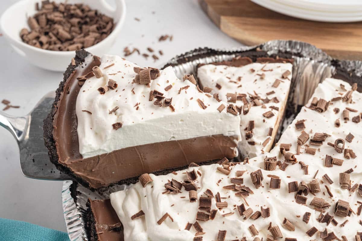 A no bake chocolate cream pie with a slice being lifted, showing layers of chocolate filling and whipped topping, Oreo crust garnished with chocolate shavings.