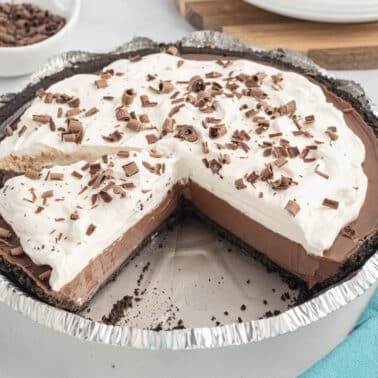 A chocolate cream pie in a foil pan with a slice missing, topped with whipped cream and chocolate shavings, sits on a white surface.