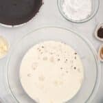A glass bowl of cream with chocolate pieces, surrounded by a chocolate crust, powdered sugar, butter, instant coffee, and vanilla on a marble surface.