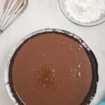 A chocolate pie with an Oreo crust sits on a marble surface next to a whisk, a bowl of powdered sugar, and a blue cloth.