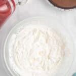 A glass bowl of whipped cream sits next to a red hand mixer with beaters and a chocolate pie in a foil pan on a marble countertop.