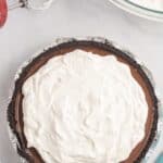 A chocolate cream pie with a whipped topping in an aluminum pie dish sits on a countertop next to a bowl of whipped cream and a hand mixer.