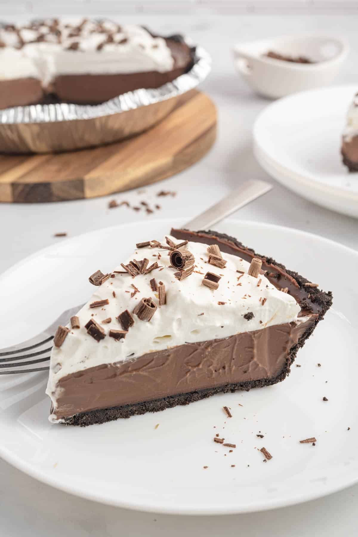 A slice of chocolate cream pie with whipped topping, Oreo crust, and chocolate shavings sits on a white plate with a fork; the rest of the pie is in the background.