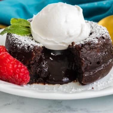A chocolate lava cake with a molten center, topped with whipped cream and a mint leaf, served with raspberries and a dusting of powdered sugar on a white plate.
