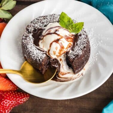 Chocolate lava cake with a scoop of vanilla ice cream, garnished with mint and powdered sugar, served on a white plate with sliced strawberries and a gold spoon.