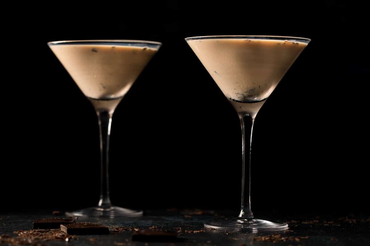 Two martini glasses filled with a chocolate martini are placed on a dark surface, with chocolate pieces and shavings nearby against a black background.
