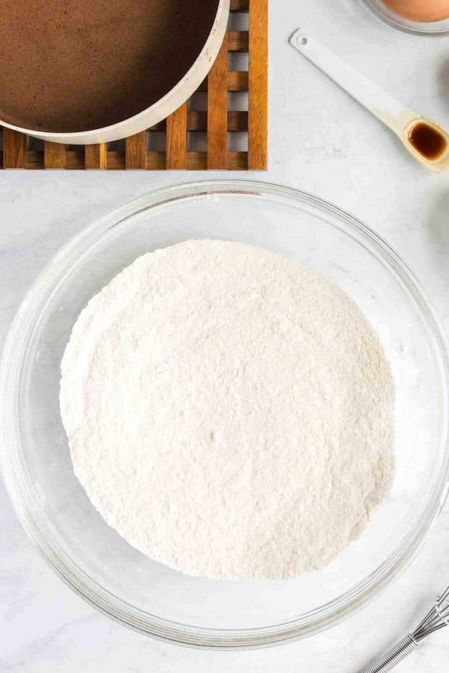 Baking ingredients on a counter including a bowl of flour mixture, a saucepan with chocolate, two eggs, a whisk, vanilla extract, baking soda, and a cup of milk.