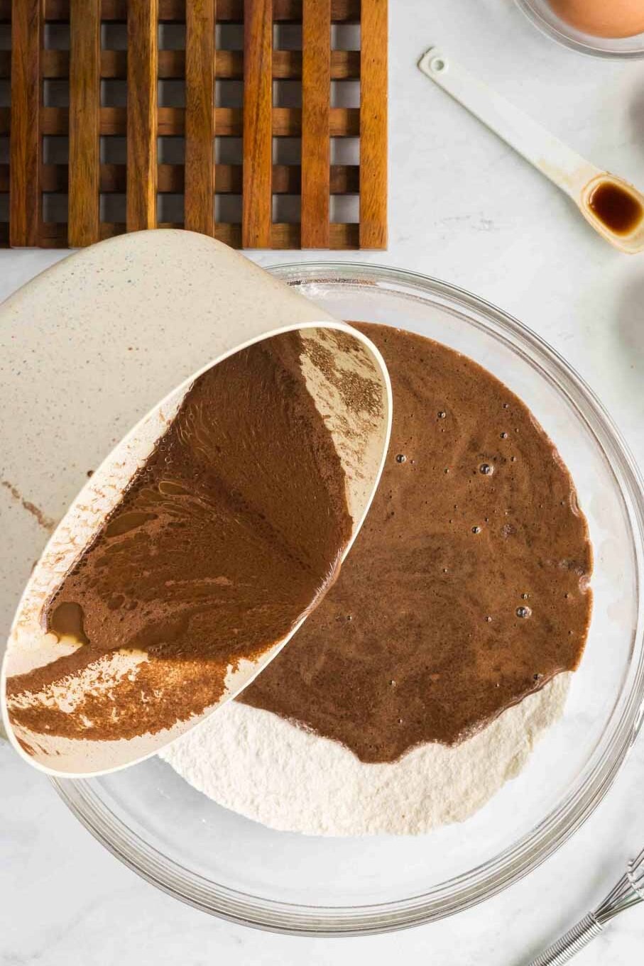 A saucepan pours chocolate mixture into a bowl of flour; next to it are eggs, vanilla, salt, milk, a whisk, and a wooden trivet on a marble surface.