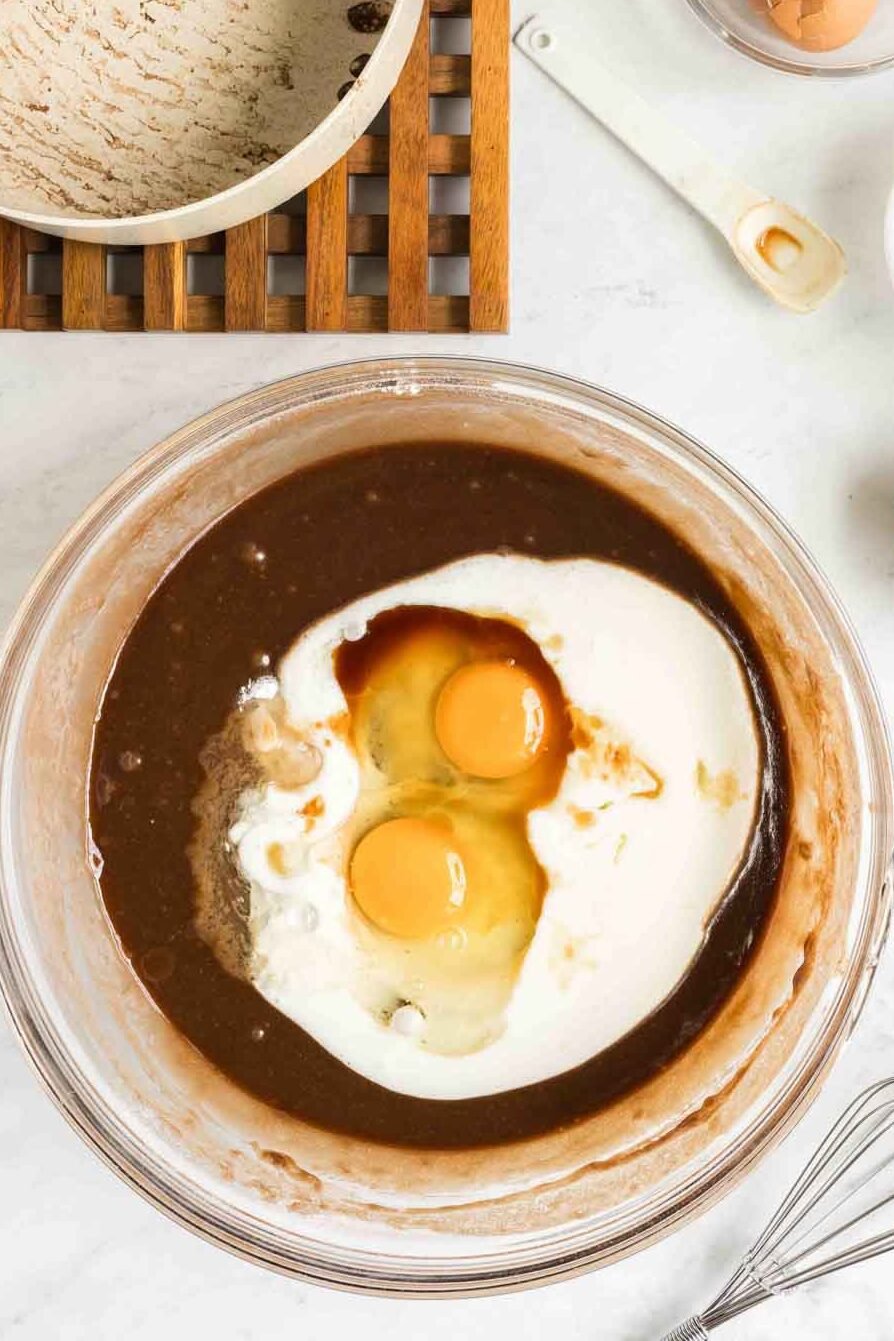 A mixing bowl with eggs, milk, and vanilla atop a brown batter, next to a bowl of flour, eggshells, a whisk, and measuring spoons on a white countertop.
