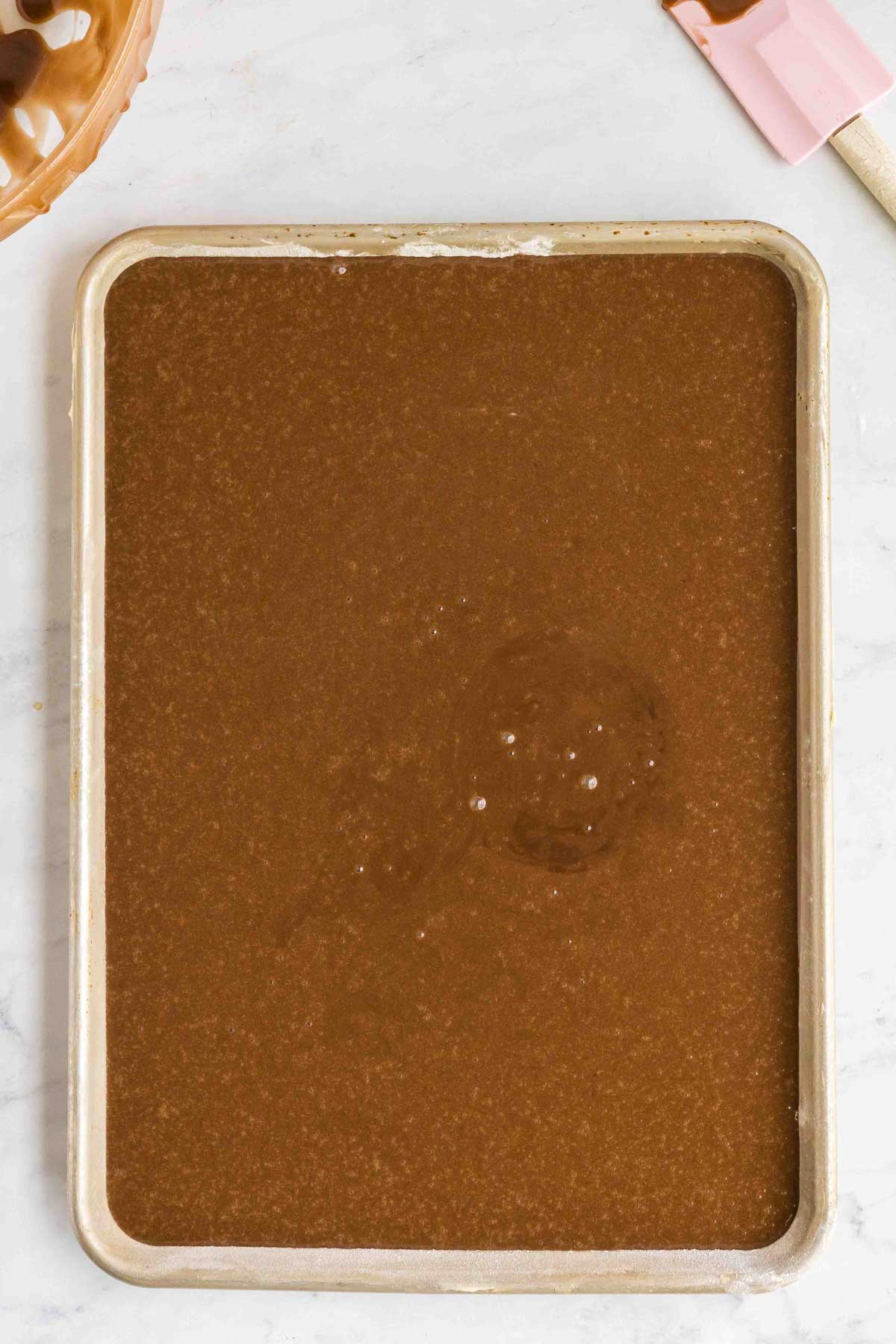 A baking sheet filled with an even layer of liquid chocolate mixture sits on a white marble surface next to a pink spatula.