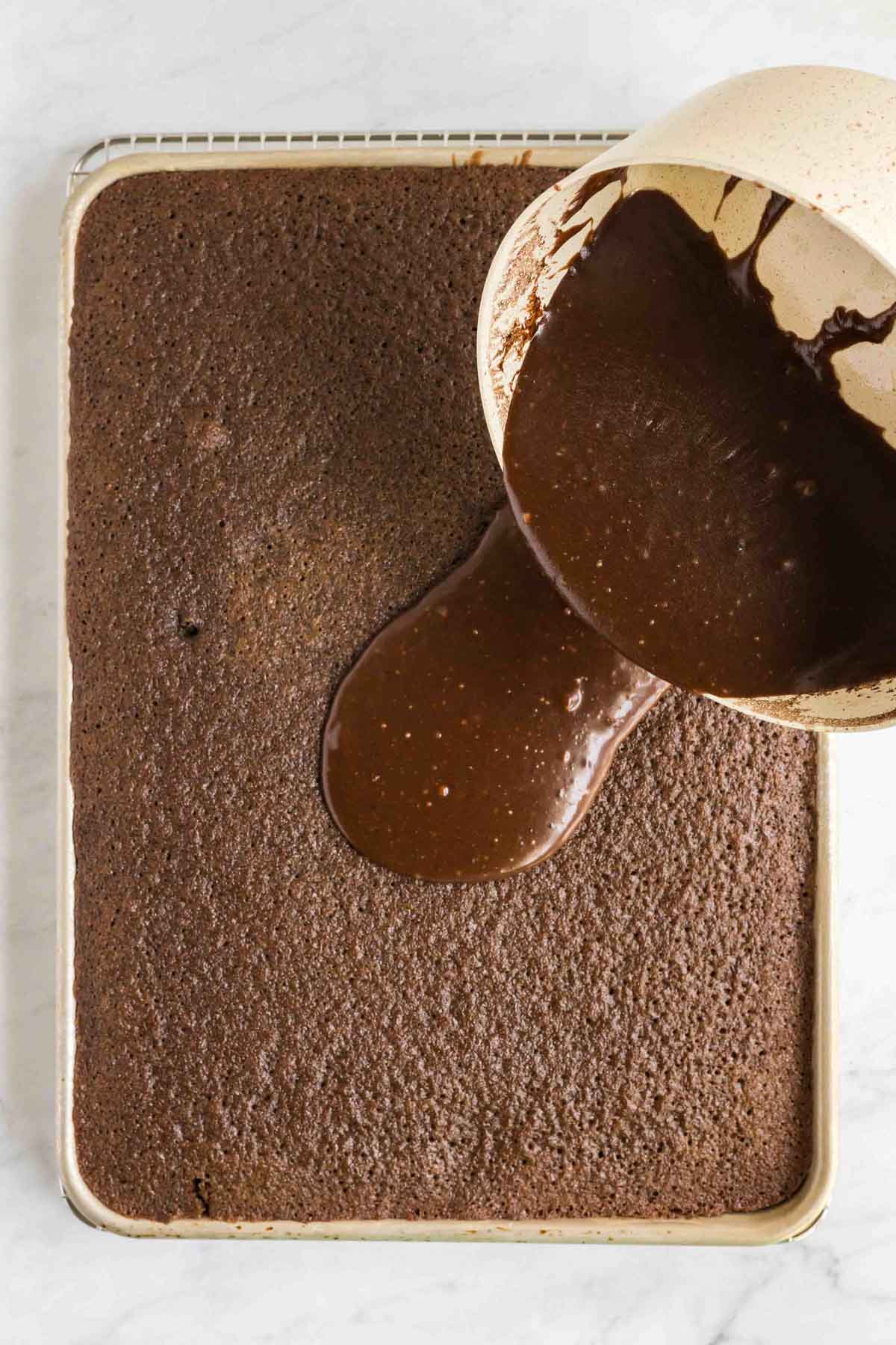 A large chocolate sheet cake on a baking tray with chocolate icing being poured onto it from a bowl.