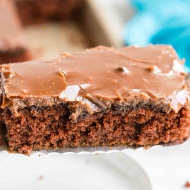 A close-up of a spatula holding a slice of chocolate sheet cake with chocolate frosting.