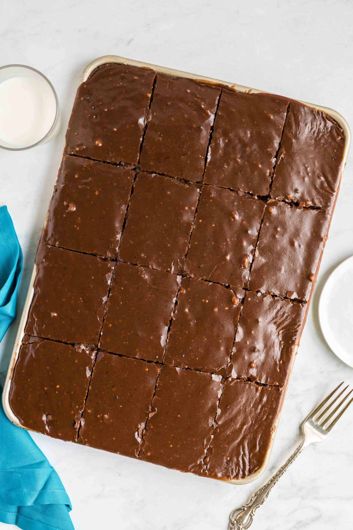 A rectangular tray of chocolate sheet cake cut into sixteen even squares, placed on a white surface with a fork, napkin, and glass nearby.