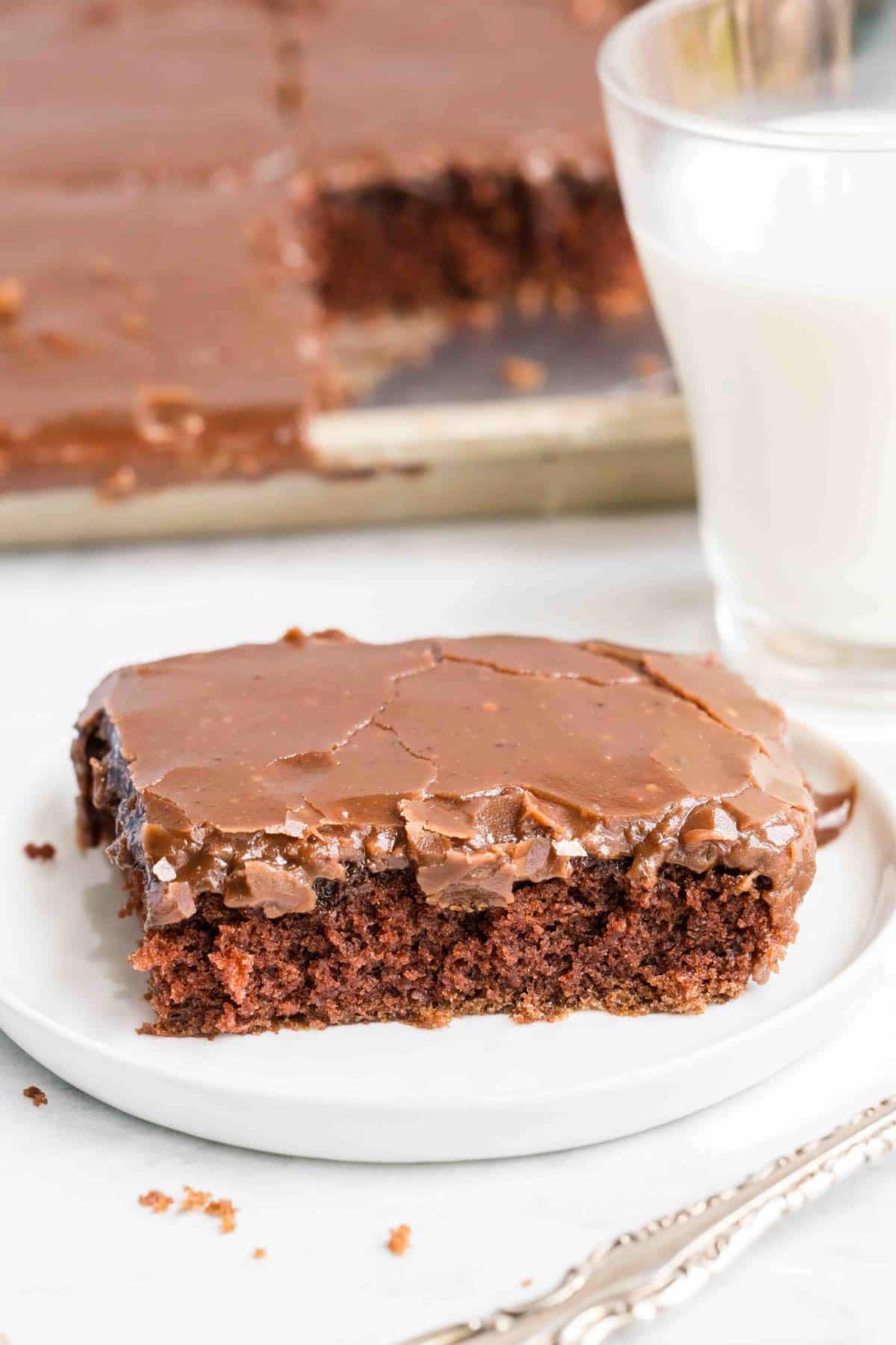 A square piece of chocolate sheet cake with chocolate icing on a white plate, next to a glass of milk.
