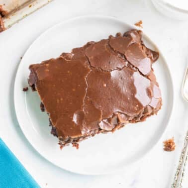 A piece of chocolate sheet cake with chocolate icing on a white plate, next to a silver fork and a blue napkin.