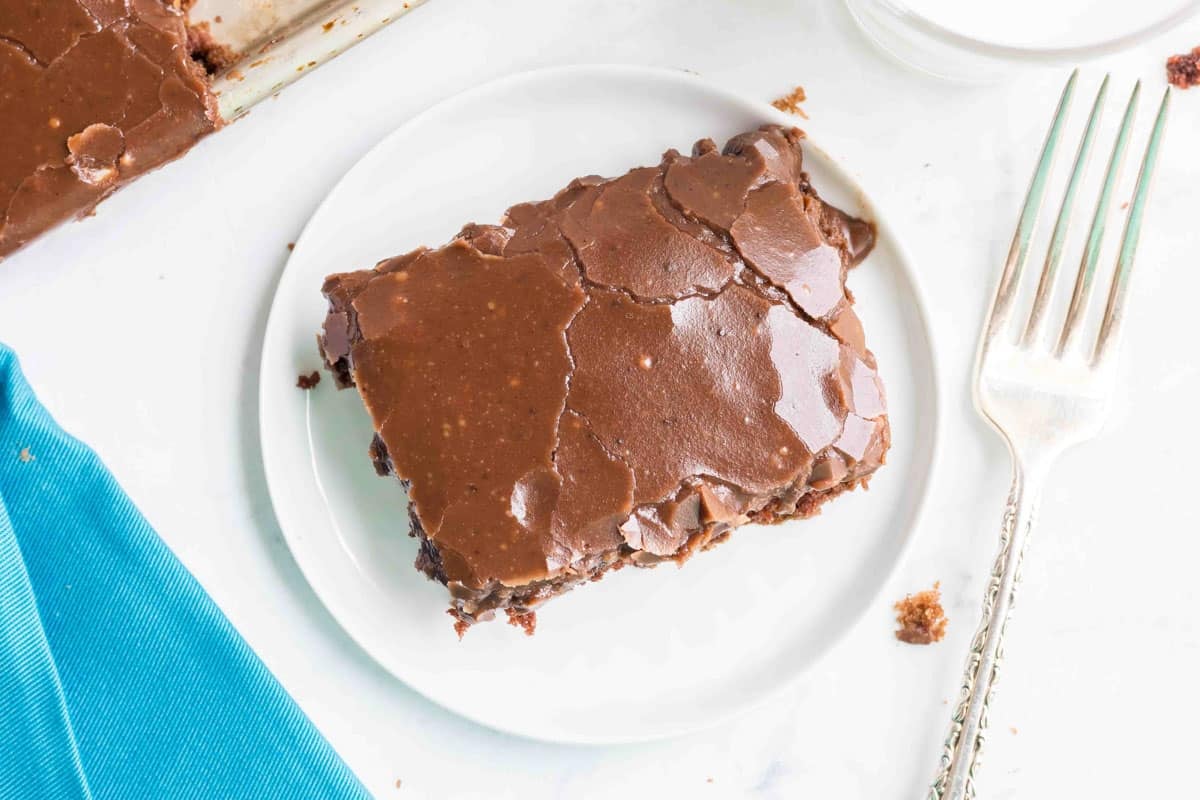 A piece of chocolate sheet cake with chocolate icing on a white plate, next to a silver fork and a blue napkin.