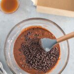 A glass bowl with brownie batter and chocolate chips being mixed with a spatula, next to a parchment-lined baking pan and a small bowl of liquid.