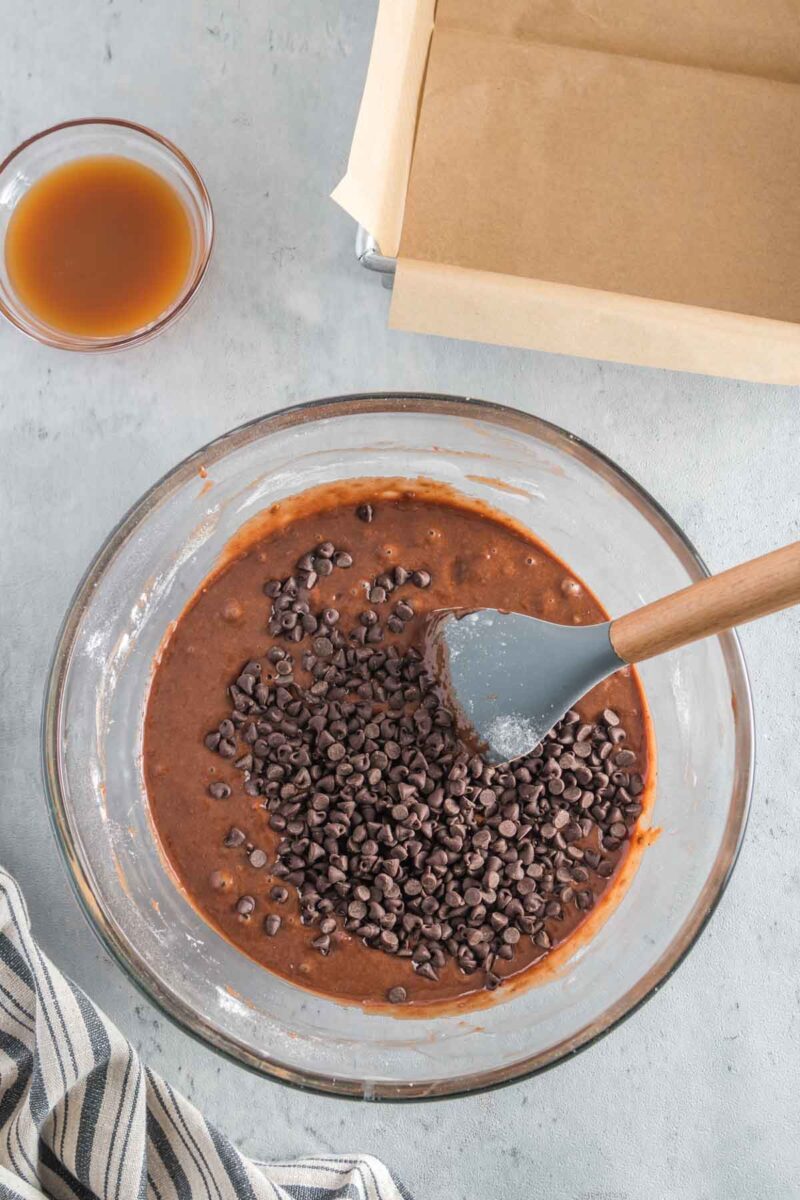 A glass bowl with brownie batter and chocolate chips being mixed with a spatula, next to a parchment-lined baking pan and a small bowl of liquid.