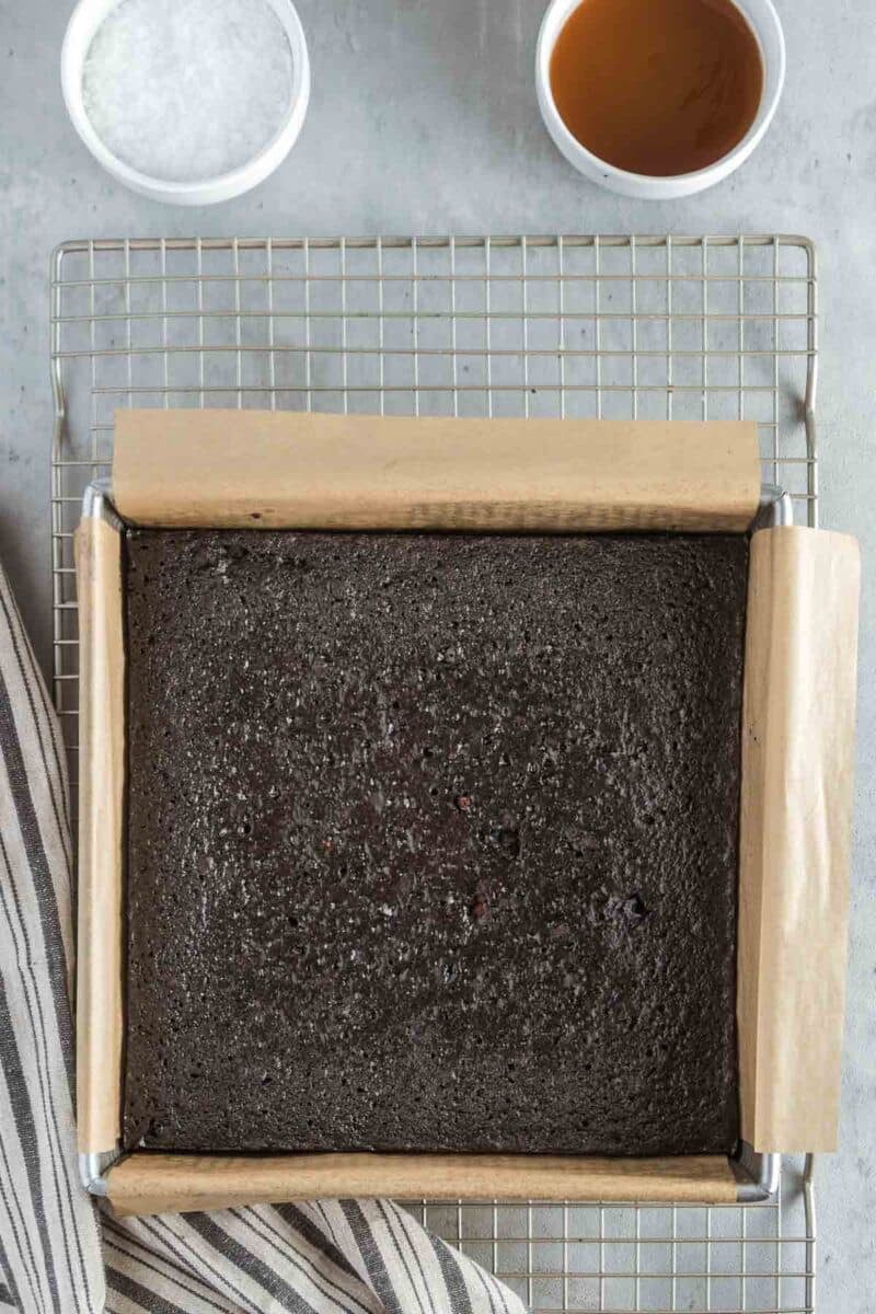 A square pan of baked brownies lined with parchment paper on a wire rack, with a bowl of salt and a cup of brown liquid above the pan.