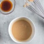 A bowl of light brown batter, a glass dish with dark liquid, and a metal whisk on a light gray surface with a striped cloth in the corner.