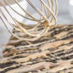 A whisk drizzling light brown glaze over a rectangular chocolate cake.