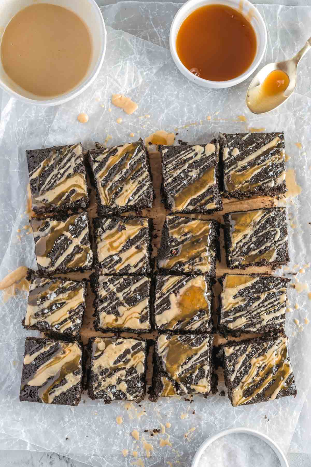 Sixteen espresso brownies are arranged in a grid on parchment paper, drizzled with light brown and caramel sauces. Two bowls with additional sauces are placed nearby.