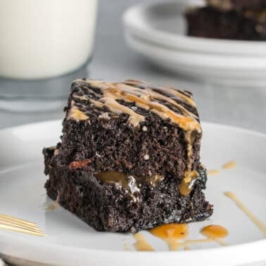 A square piece of espresso brownies with caramel drizzle sits on a white plate, with a glass of milk and more cake slices in the background.