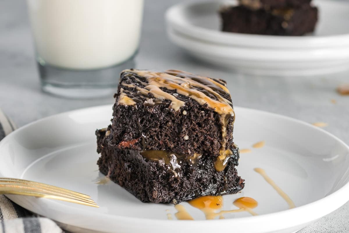A square piece of espresso brownies with caramel drizzle sits on a white plate, with a glass of milk and more cake slices in the background.