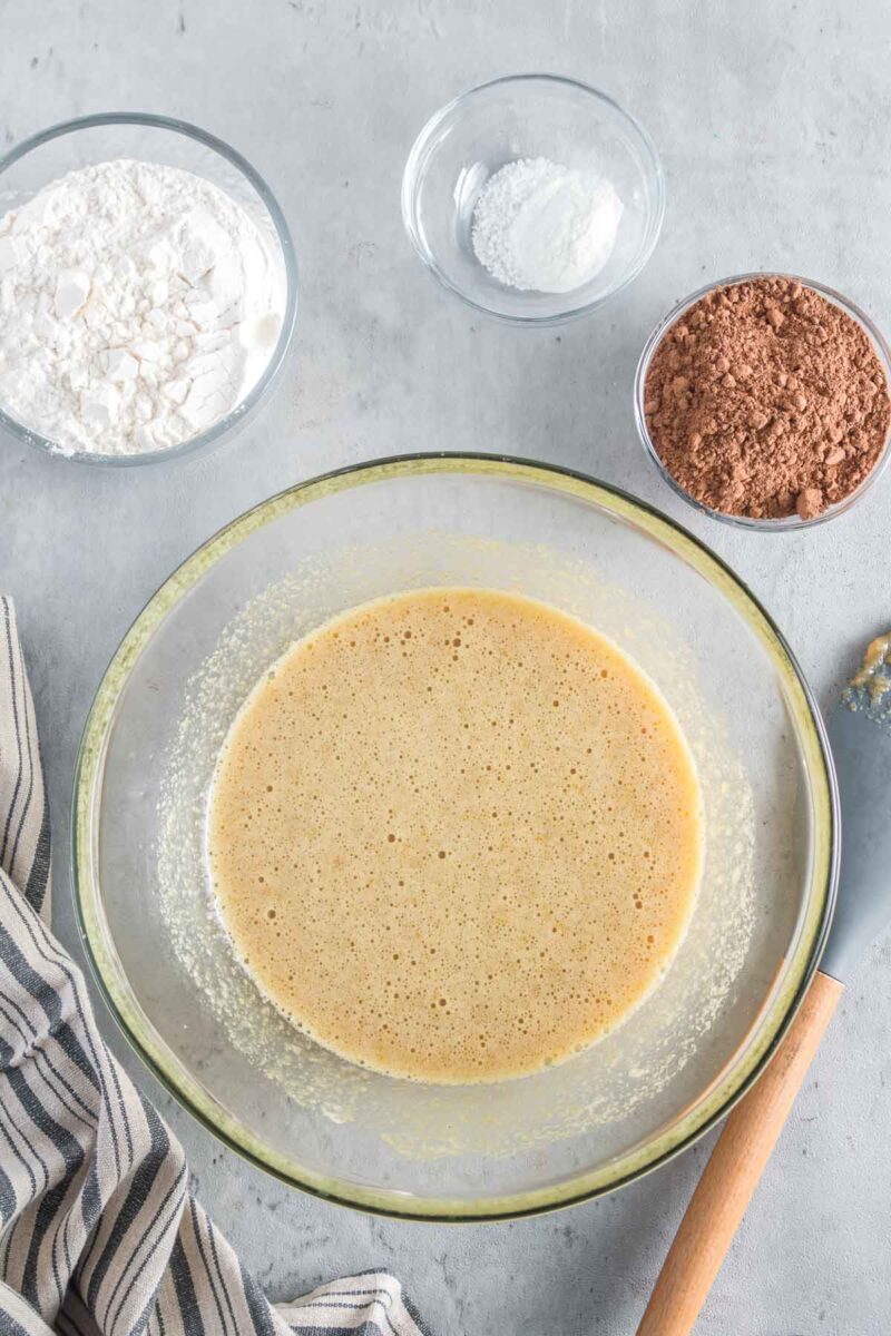 A glass bowl with a whisked yellow batter, surrounded by bowls of flour, cocoa powder, and baking powder on a gray surface. A striped cloth and spatula are beside the bowl.