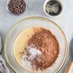 A glass bowl with batter, cocoa powder, and baking powder, next to a bowl of chocolate chips and a cup of coffee on a gray surface.
