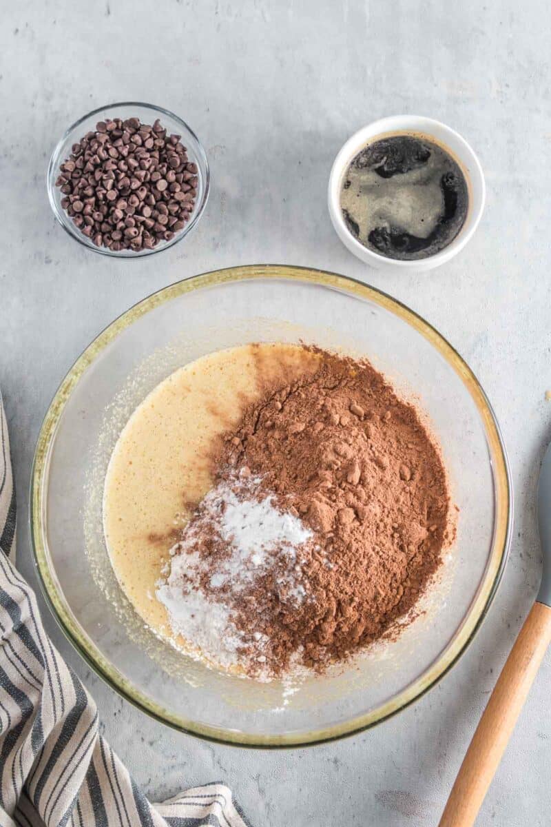 A glass bowl with batter, cocoa powder, and baking powder, next to a bowl of chocolate chips and a cup of coffee on a gray surface.